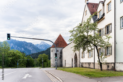 Fototapeta Naklejka Na Ścianę i Meble -  A historic tower and gate stand at the entrance of a European village road, surrounded by white buildings and green trees.
