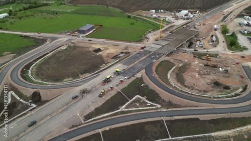 Aerial flyover footage of construction worksite with workers and machinery building a bridge. High angle shot of highway overpass being built on an industrial location. Establishing shot.
