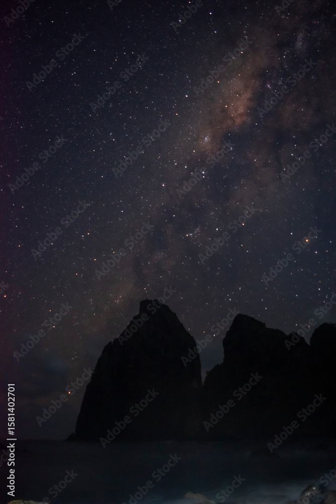 Fototapeta premium Silhouette of coastal rocks beneath a star-filled sky at a hidden beach, with the Milky Way glowing above the dark sea horizon.