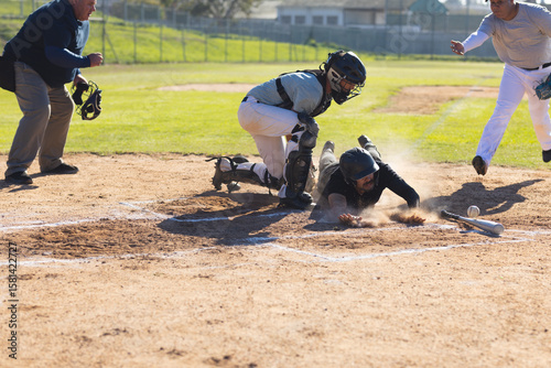 Male baseball players and umpire sliding and tagging at home plate with catcher's mitt and baseball