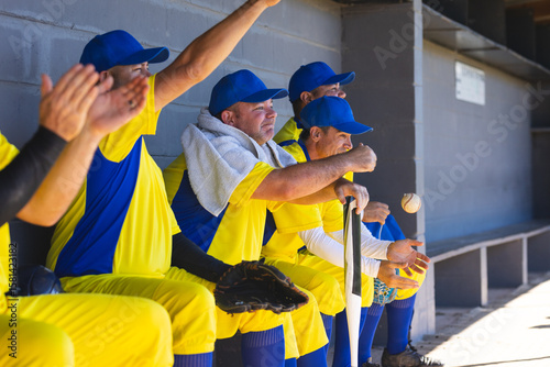 Male baseball teammates cheering and passing baseball with bat, glove, towel on dugout bench