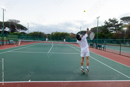 Asian male tennis player serving ball with racket over net, rival awaiting on hardcourt, copy space