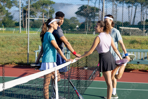 Diverse tennis players in sportswear leaning over net on park court holding rackets and chatting