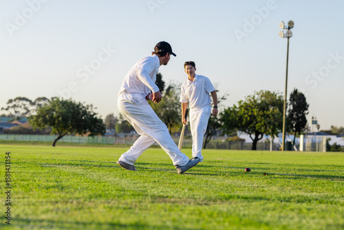 Two male teammates in white uniforms moving to stop cricket ball at suburban ground with bat