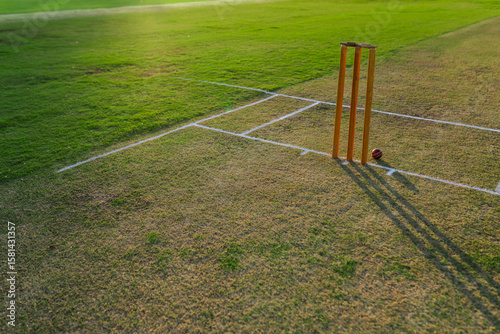 Wooden cricket stumps standing on worn grass pitch with red ball resting under sharp shadows