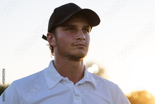 Man gazing across golf course at sunset, wearing black baseball cap and white polo shirt