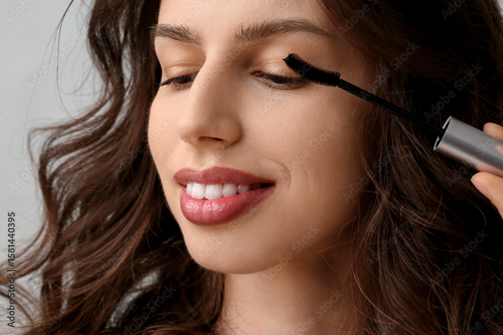 Beautiful young woman applying mascara on grey background, closeup Beautiful young woman applying mascara on grey background, closeup