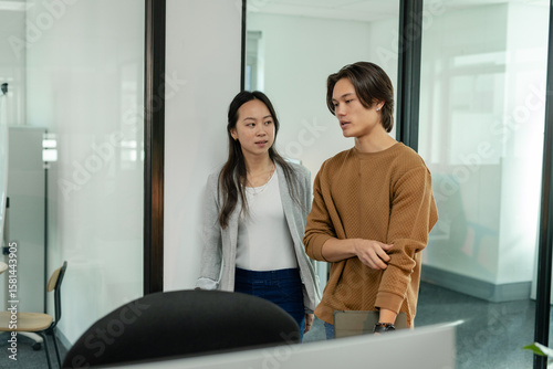 Asian coworkers in business attire standing at office desk reviewing content on computer monitor