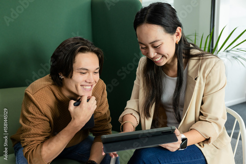 Asian colleagues sitting on green cushioned seating at office lounge, sharing tablet and smartphone