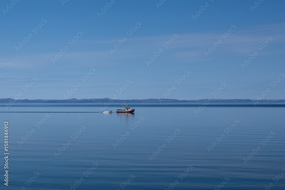 Fototapeta premium A small fishing boat glides across the calm, blue ocean, with a colorful blue sky. There are thin clouds over a mountain landmark. The red fishing boat is towing a small white open rowboat or skiff.