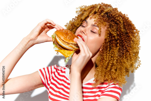 Joyful woman enjoying a delicious hamburger in a vibrant striped shirt, showcasing excitement and a love for fast food against a bright white background