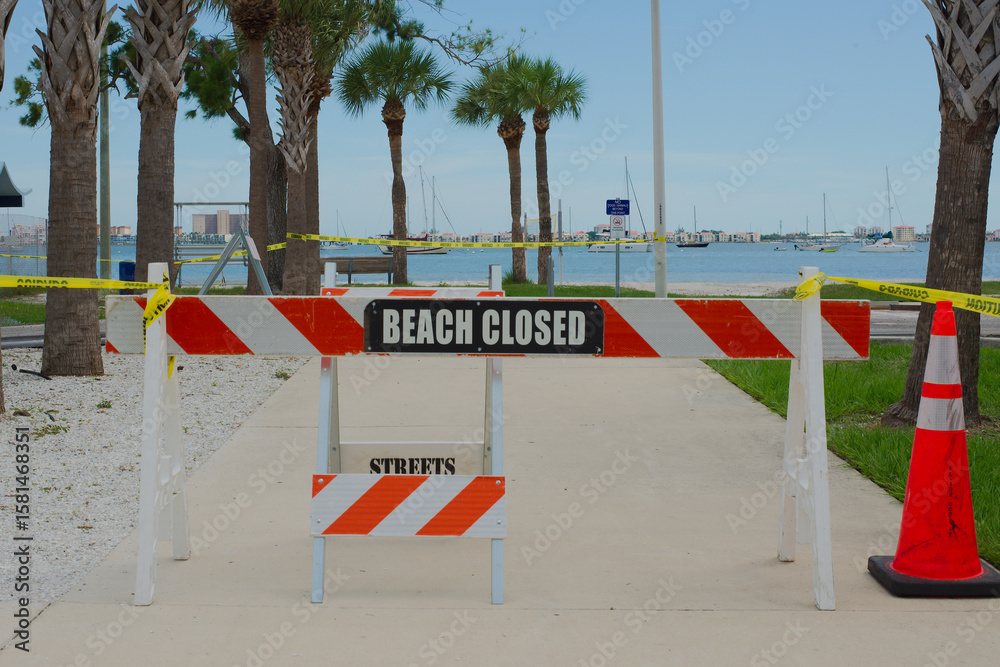 Obraz premium Beach Closed Sign on Walkway with Caution Tape and Palm Trees in Gulfport, Florida. Barricade blocking access to a beach walkway with a 'Beach Closed' sign, surrounded by palm trees and caution tape,