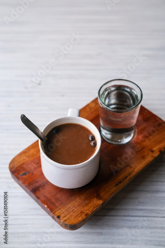 Steaming cup of kopi susu served with a glass of water on a rustic wooden tray at a cozy local coffee shop. Warung kopi Pontianak.