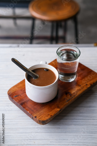 Steaming cup of kopi susu served with a glass of water on a rustic wooden tray at a cozy local coffee shop. Warung kopi Pontianak.
