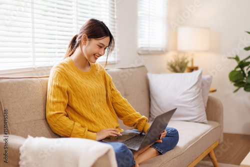 Excited happy young asian woman using laptop sitting on sofa at home
