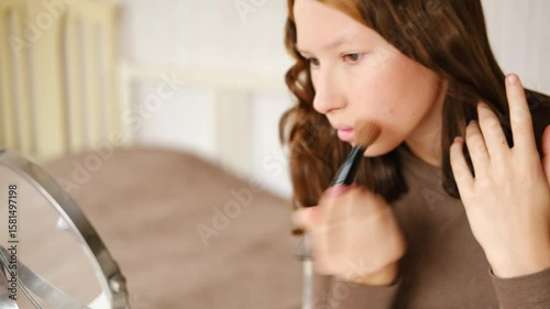 Teen Girl putting on makeup looking at mirror. Beautiful Teenager caring her face. 14 years old female applied cosmetics. Natural beauty concept. Skin care, make-up. Daily make up. Beautiful Teenage