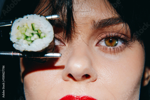 Close-up portrait of a woman holding sushi with chopsticks, showcasing vibrant eyes and bold red lips, perfect for food and culture themes.