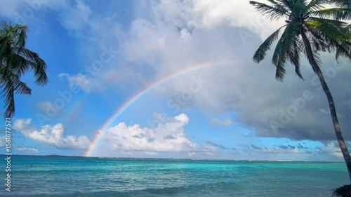 A stunning rainbow arches over a tranquil turquoise ocean with lush palm trees. A beautiful, serene and peaceful tropical paradise scene.