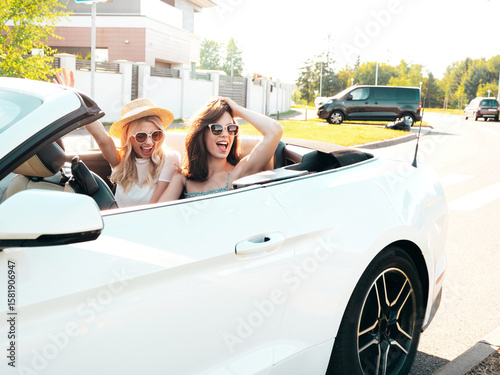 Portrait of two young beautiful and smiling hipster female in convertible car. Sexy carefree women driving. Positive models riding and having fun in sunglasses outdoors. Enjoying summer days