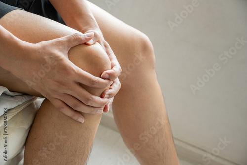 An Asian man seated on a bed clutches his right knee, showing clear signs of discomfort. A soft indoor setting enhances the medical focus on joint pain. Healthcare and medical concept. Copy space.