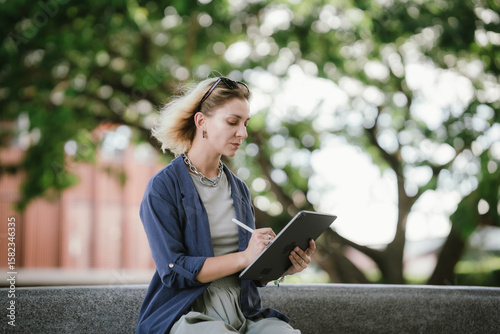 A confident woman uses a tablet and stylus outdoors, creating digital art with focus and style, embodying modern creativity, technology, and freelance lifestyle in an urban setting.