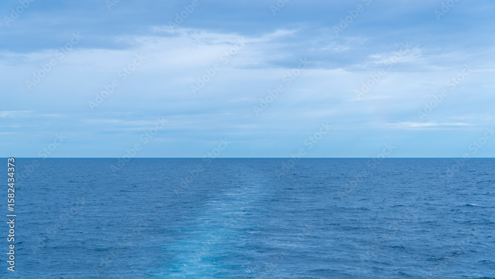 Fototapeta premium Blue ocean with a ship's wake trailing towards the horizon under a cloudy sky