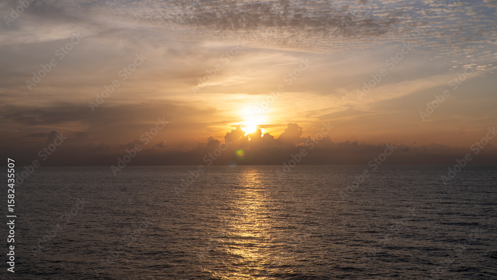 Fototapeta premium Sunrise over the ocean with the sun partially obscured by clouds, casting a golden reflection