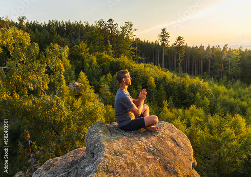 Young man meditating, praying on top of rock with trees behind. Active lifestyle, mental health background