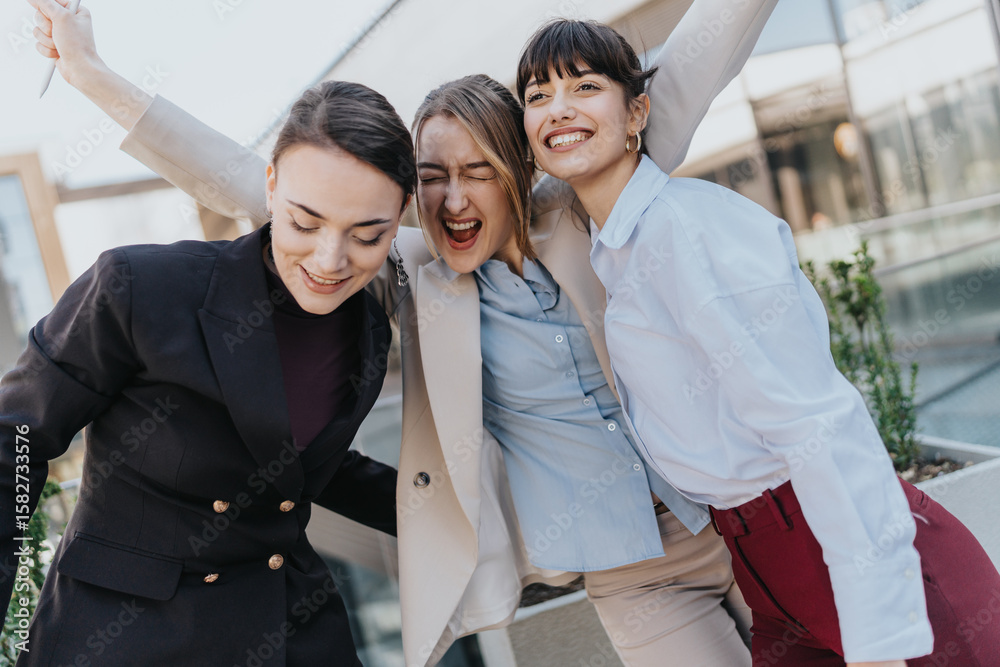 Obraz premium A happy trio of young professional women expressing joy and camaraderie in an outdoor modern setting. A moment capturing success and teamwork in a professional and urban background.