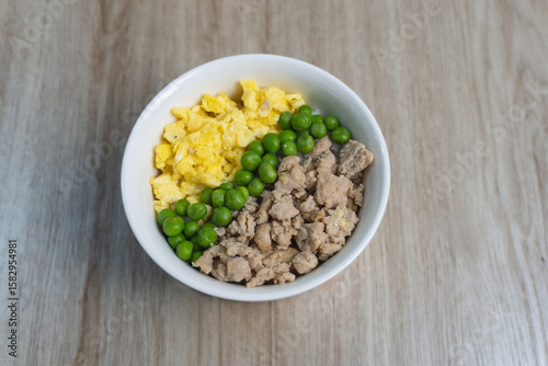 Top-down view of a simple and balanced meal in a white bowl, featuring scrambled eggs, green peas, and ground chicken, placed on a wooden table