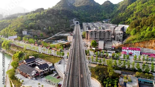 Aerial view of high-speed railway tracks traversing mountainous terrain with surrounding developments in a serene landscape