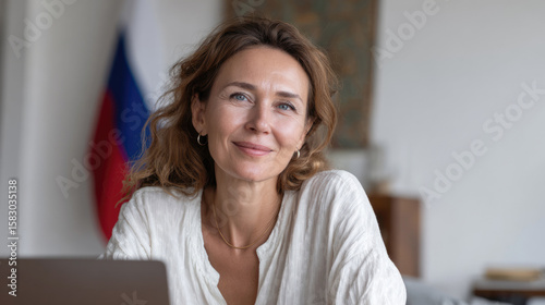 A Russian language teacher at a table with a laptop, against the background of the Russian Federation flag, preparing for an online lesson.