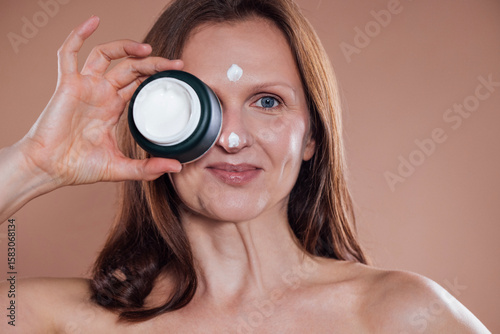 Obraz na plátne A middle-aged woman holds a jar of cream to her face and smiles