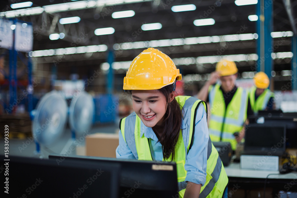 Fototapeta premium Smiling industrial worker in protective helmet using desktop computer in distribution center, focusing on inventory management tasks.
