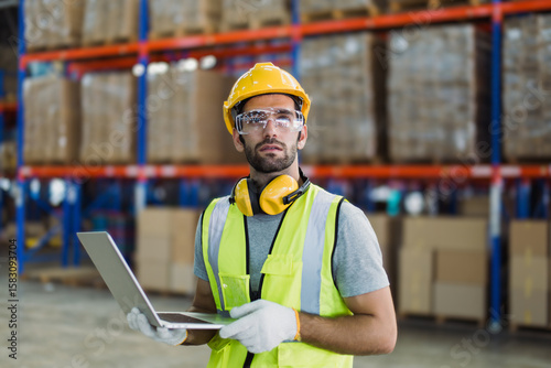 Cuadro en lienzo Warehouse logistics coordinator wearing safety gear using laptop to manage transportation schedules and cargo operations in a busy industrial distribution center