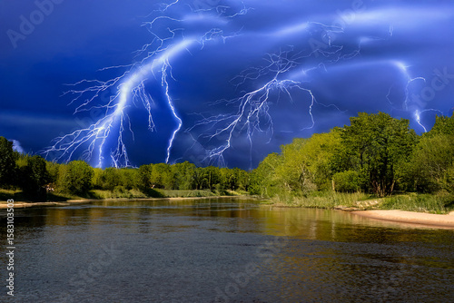 Multiple bright lightning bolts streak across a vivid blue storm sky above a peaceful river surrounded by lush green forest.