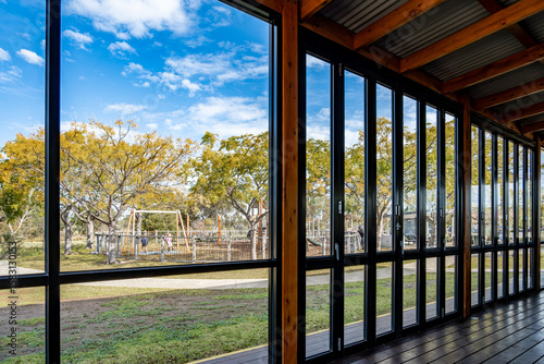 Canvas-taulu An enclosed patio or covered outdoor seating area with bi-fold glass doors with a view of sunny suburban park with trees and a children's playground outside