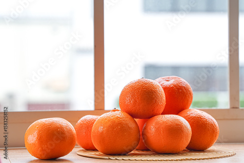 A stack of tangerines sits on a woven mat in front of a window, bathed in natural light.