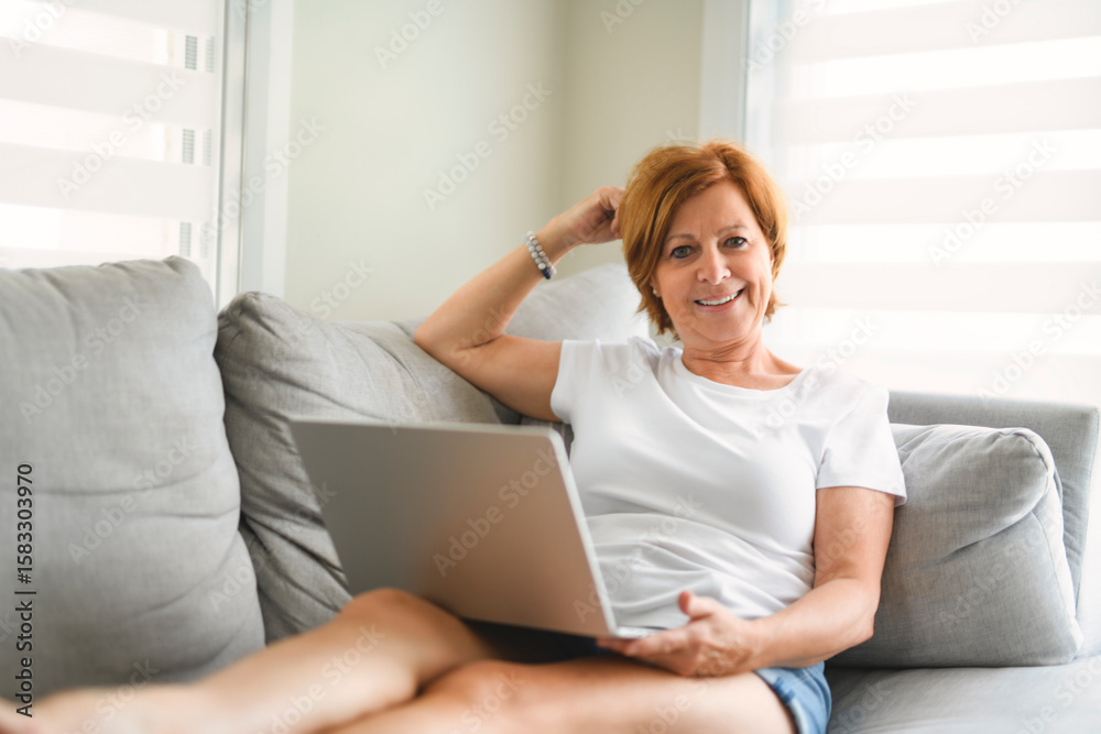 Happy modern European senior woman using laptop, touching eyeglasses and smiling to camera, enjoying digital leisure, sitting on couch Happy modern European senior woman using laptop, touching eyeglasses and smiling to camera, enjoying digital leisure, sitting on couch