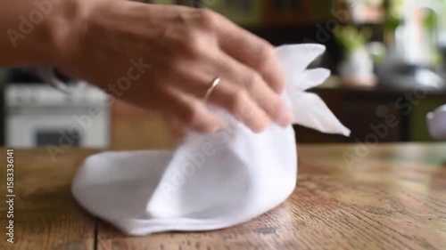 Napkin folding. Hands fold a white cloth napkin atop a wooden table. A wedding ring adorns one finger. Soft, natural light