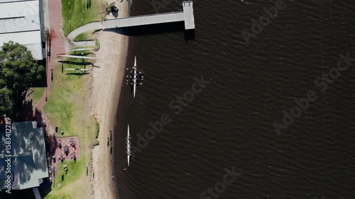 Aerial view of groups of rowers practice on serene water close to a wooden dock with people relaxing on the sandy bank