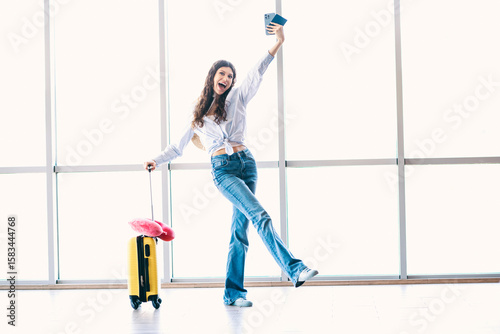 Cheerful young woman with suitcase and passport celebrating travel at airport, ready for vacation or journey