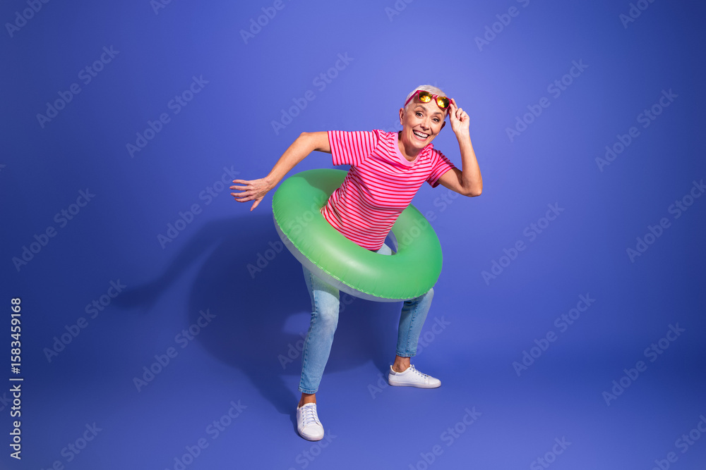 Obraz premium Joyful mature woman in casual attire with green swimming ring on violet background, showcasing a playful summer vibe