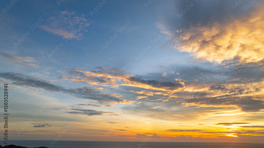 Fototapeta premium A breathtaking sunset paints the sky in vivid hues of orange and blue over a coastal town, with lush green hills framing the scene. A distant hilltop structure adds a serene tranquil landscape.