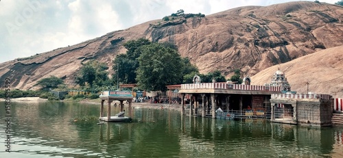 A scenic view of Saravana Poigai, the sacred temple tank at Thiruparankundram, Tamil Nadu, surrounded by rocky hills and lush trees, adjacent to the revered Thiruparankundram Murugan Temple