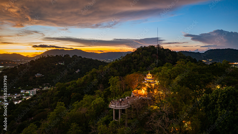 Fototapeta premium A scenic aerial view of a town nestled among lush green mountains at sunset, with a tower structure perched on the hillside and a vibrant orange sky creating a warm, tranquil atmosphere.