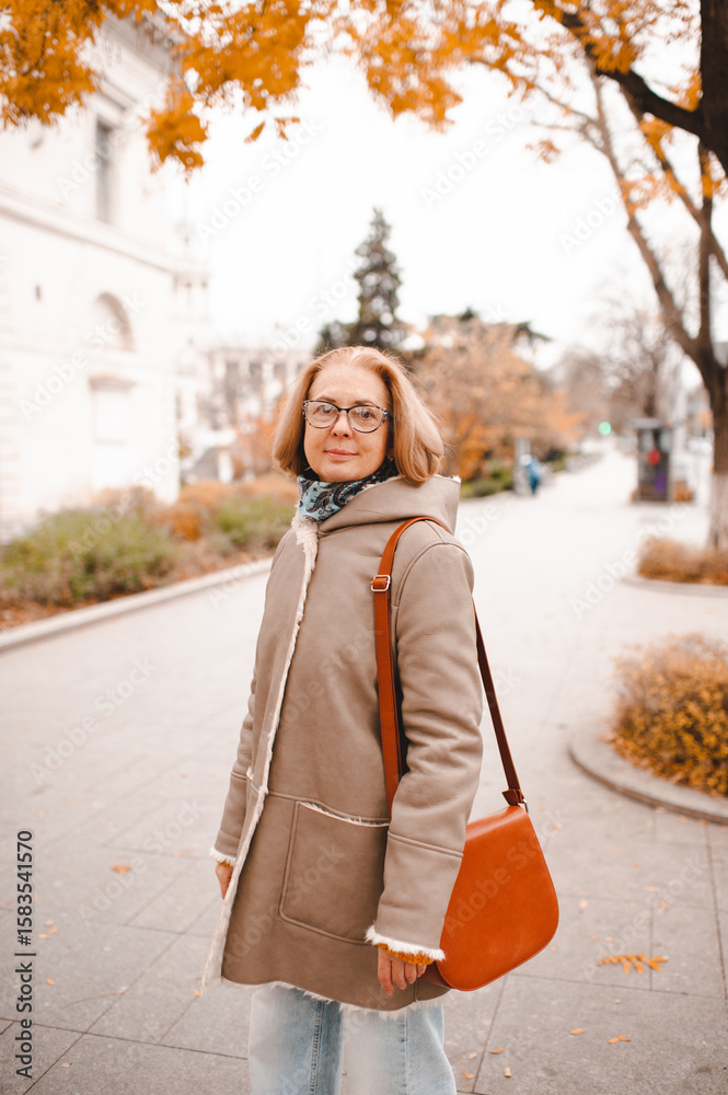 Fototapeta premium Smiling mature woman wearing eye glasses and autumn jacket over yellow leaves outdoors. Fall season. Senior adult lifestyle.