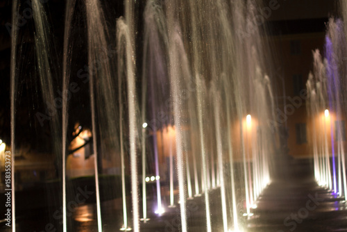 Vibrant streams of water ascend from a modern fountain, brightly lit against the dark night sky, creating a captivating urban display in Reggio Emilia