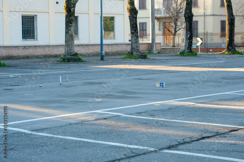 An empty, asphalt urban parking lot with white lines, trees, and buildings in the background, showing some scattered litter on the ground.