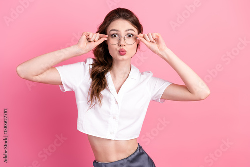 Charming young woman wearing a white shirt makes a playful gesture with glasses standing against a pink background